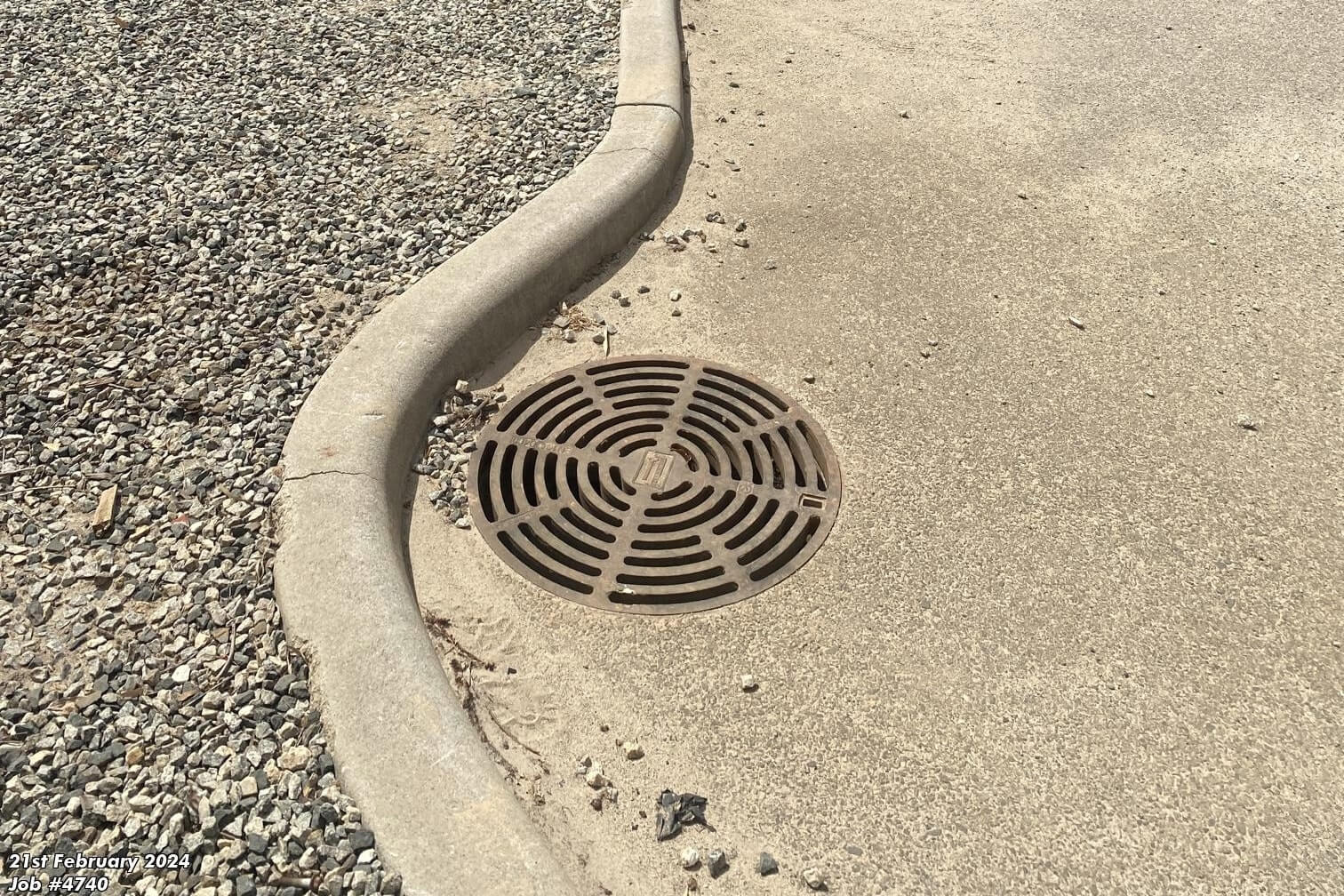 Commercial stormwater drain grate installed in a paved area next to a kerb and gravel landscaping