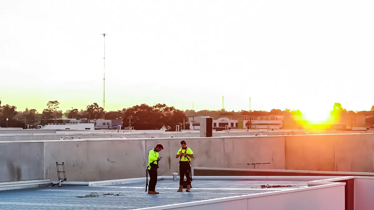Two tradespeople in hi-vis inspecting a large commercial metal roof at sunset